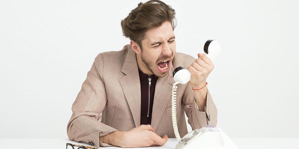 Ein Mann sitzt am Tisch und schreit wütend in den Hörer eines weißen Festnetztelefons; daneben liegt eine Brille. Ein Mann sitzt am Tisch und schreit wütend in den Hörer eines weißen Festnetztelefons; daneben liegt eine Brille.