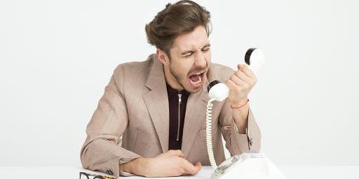 Ein Mann sitzt am Tisch und schreit wütend in den Hörer eines weißen Festnetztelefons; daneben liegt eine Brille. Ein Mann sitzt am Tisch und schreit wütend in den Hörer eines weißen Festnetztelefons; daneben liegt eine Brille.